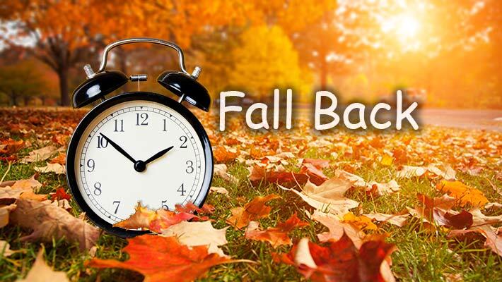 Alarm clock sitting on the ground with leaves and the text Fall Back.